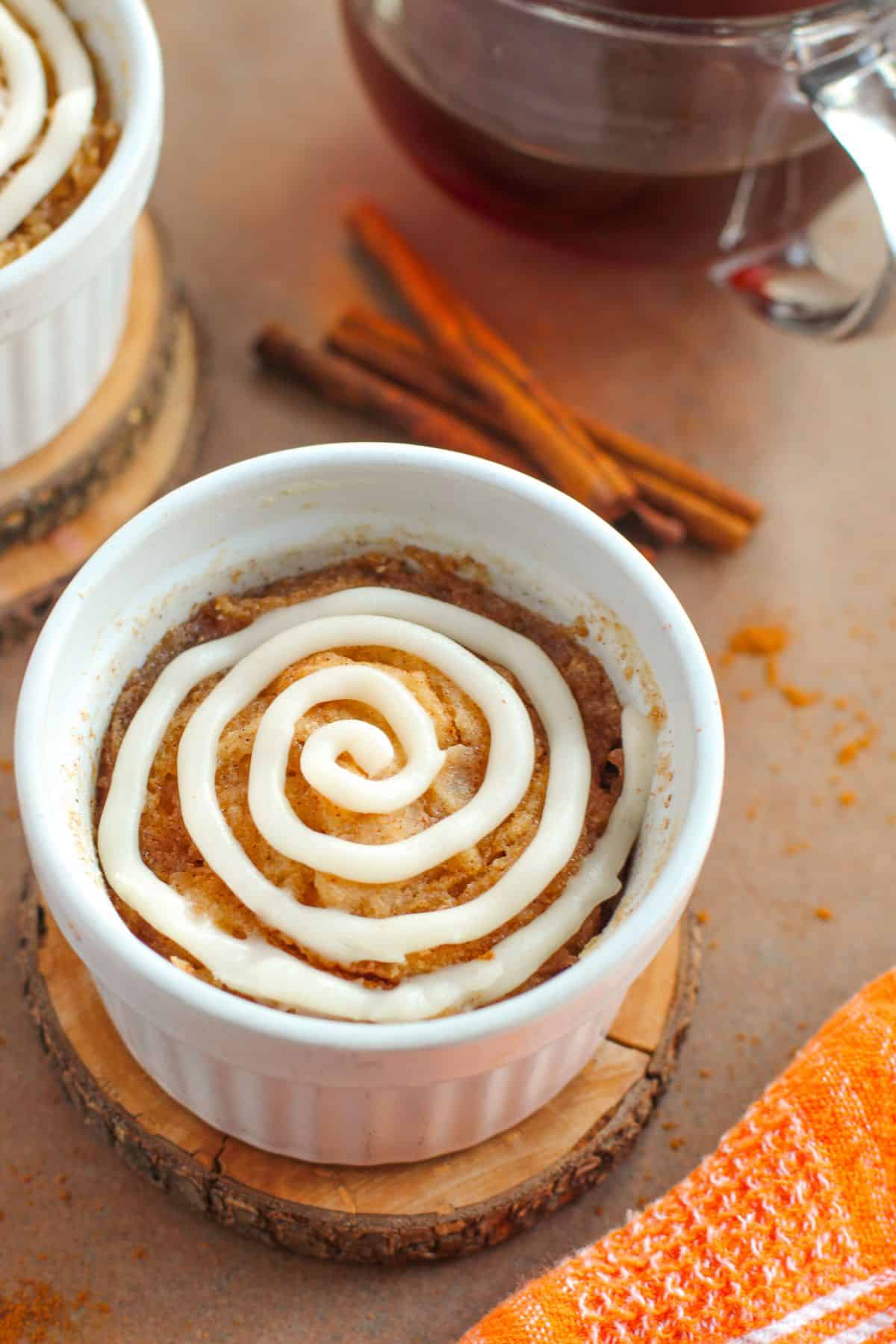 A single-serving dessert in a white ramekin, topped with a spiral of white icing, sits on a wooden coaster. Cinnamon sticks and a cup of tea are in the background.