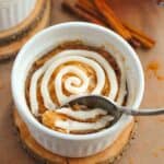 A spoon scoops into a Cinnamon Mug Cake in a white ramekin, topped with a swirl of white icing, set on a wooden coaster with cinnamon sticks in the background.