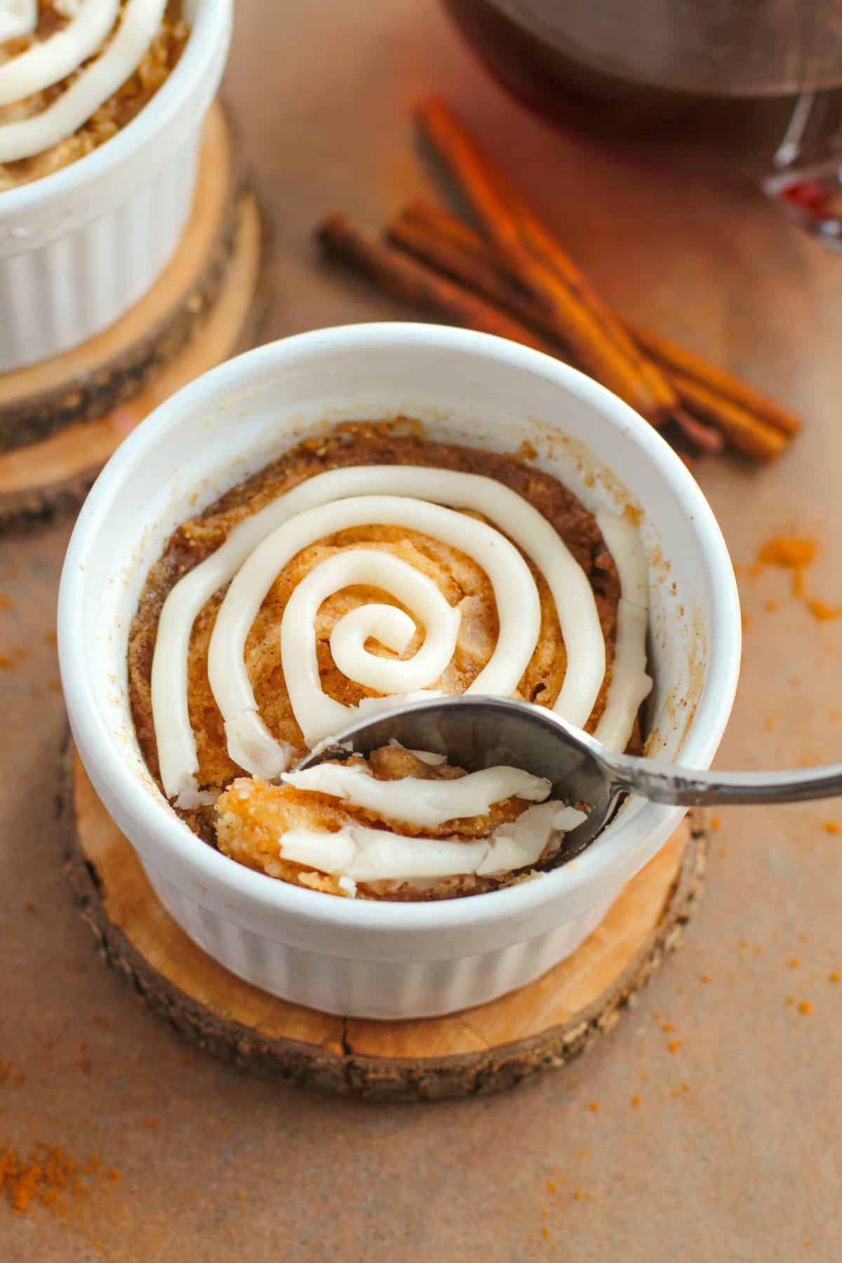 A spoon scoops into a Cinnamon Mug Cake in a white ramekin, topped with a swirl of white icing, set on a wooden coaster with cinnamon sticks in the background.