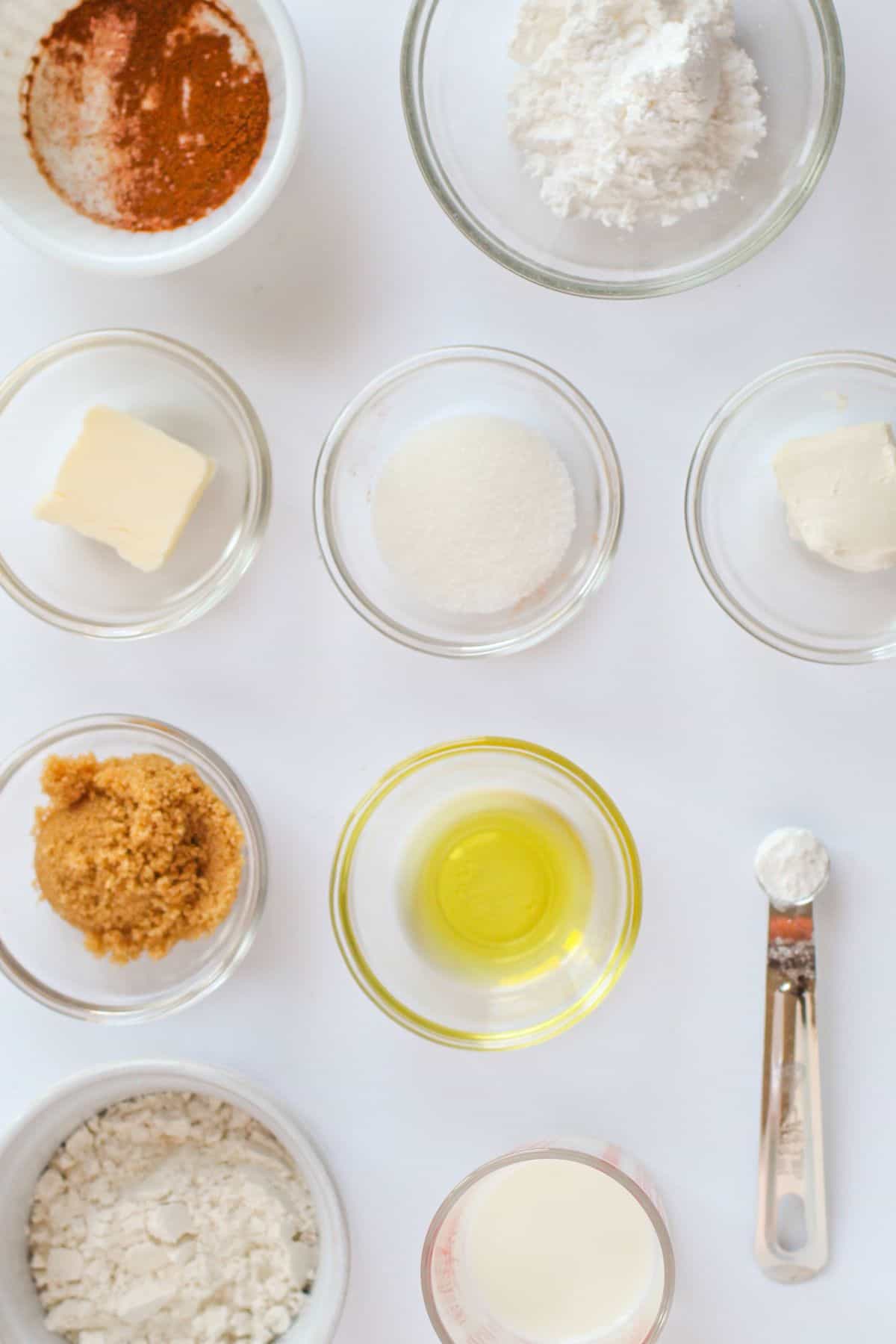 Nine small bowls containing various baking ingredients including flour, sugar, butter, oil, brown sugar, baking powder, and a measuring spoon, arranged on a white surface.