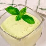 A glass cup filled with pale green smoothie, topped with three fresh basil leaves; a spoon and checkered cloth are in the background, reminiscent of the fresh flavors in Panera Green Goddess Dressing.