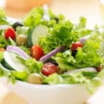 A white bowl filled with fresh salad—lettuce, cucumber slices, cherry tomatoes, red onion, black olives, and green olives—tossed in creamy Panera Green Goddess Dressing.