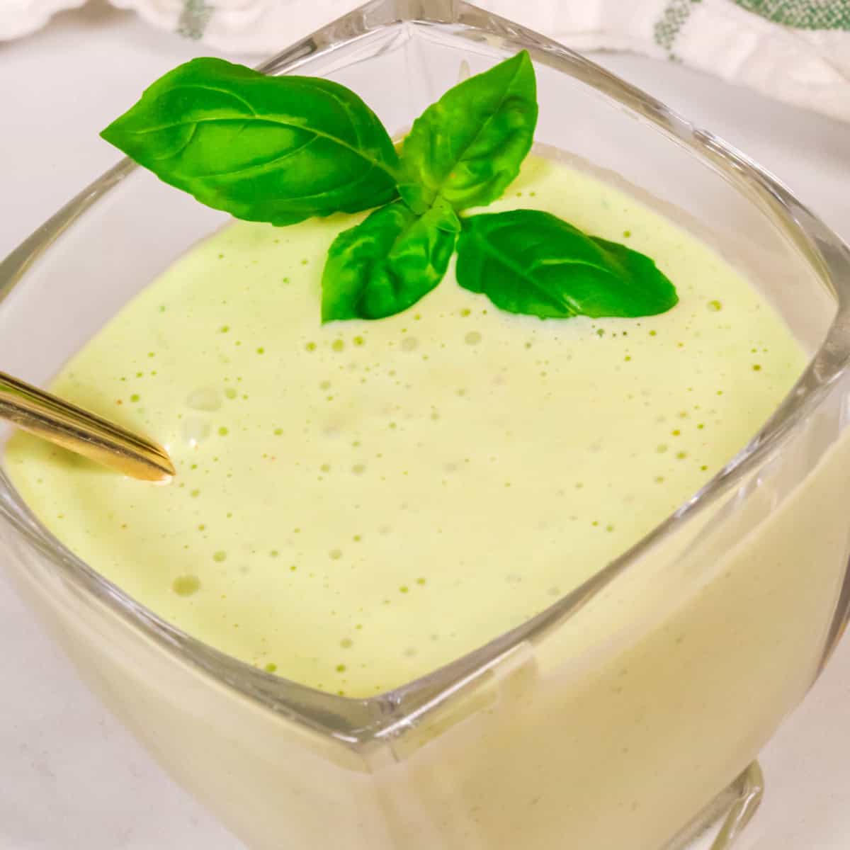 A glass of creamy green smoothie topped with fresh basil leaves, featuring a hint of Panera Green Goddess Dressing, with a gold spoon inside the glass.