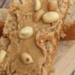 A slice of Peanut Butter Bread topped with whole peanuts sits on a wooden board, accented by a red cloth nearby.