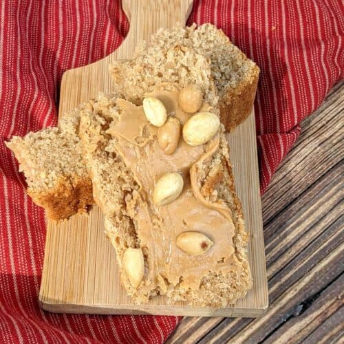 A slice topped with whole peanuts rests on a wooden board, set atop a red striped cloth on a rustic wooden surface.