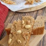 A slice of Peanut Butter Bread topped with creamy peanut butter and peanuts sits on a wooden board, with more bread and crumbs visible in the background.