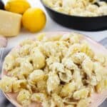 A plate of Pasta with Ricotta and Lemon is coated in a creamy cheese sauce, topped with grated cheese. A skillet with more pasta, fresh lemons, and a wedge of cheese are in the background.