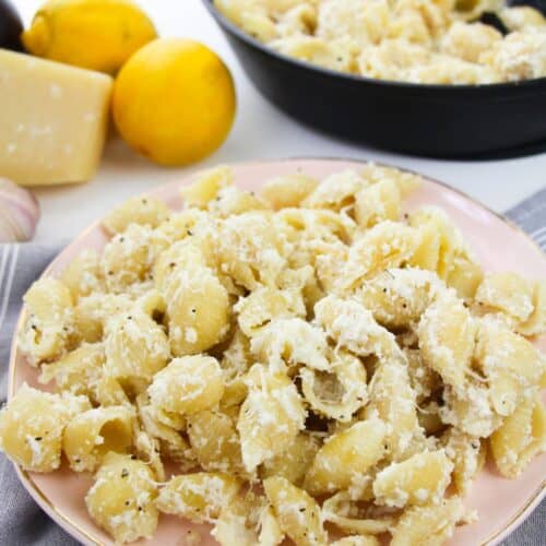 A plate of Pasta with Ricotta and Lemon is coated in a creamy cheese sauce, topped with grated cheese. A skillet with more pasta, fresh lemons, and a wedge of cheese are in the background.