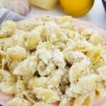 A plate of Pasta with Ricotta and Lemon, covered in a creamy white cheese sauce, sits on a striped napkin, with a garlic bulb, a lemon, and a wedge of cheese in the background.