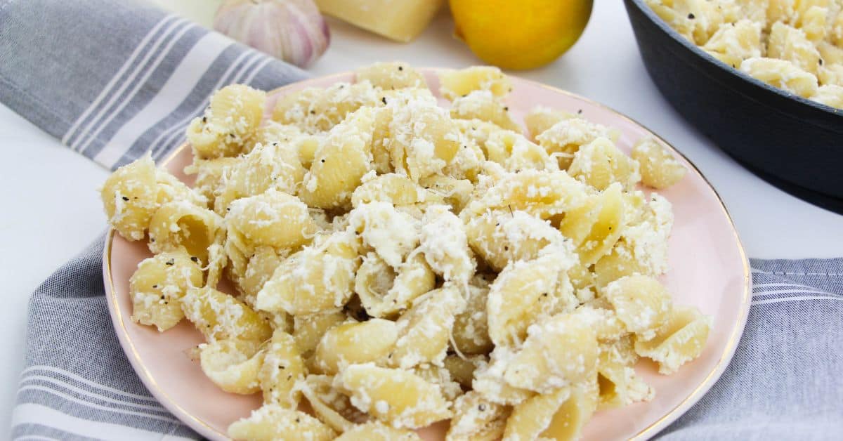 A plate of Pasta with Ricotta and Lemon, covered in a creamy white cheese sauce, sits on a striped napkin, with a garlic bulb, a lemon, and a wedge of cheese in the background.