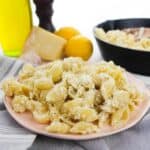 A plate of shell pasta with a creamy cheese sauce sits on a gray cloth, with a skillet, olive oil, pepper grinder, cheese, lemons, and garlic in the background—perfect for Pasta with Ricotta and Lemon lovers.