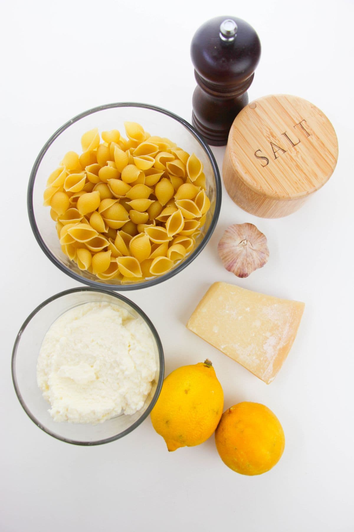 A bowl of shell pasta, ricotta cheese, two lemons, Parmesan wedge, garlic bulb, salt container, and pepper grinder on a white surface—everything you need for fresh Pasta with Ricotta and Lemon.