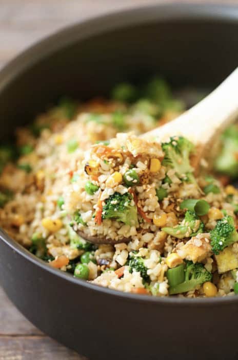 A wooden spoon scoops a vibrant mixture of fried rice, featuring broccoli, corn, peas, and carrots, from a black bowl—a colorful side dish that's perfect for serving alongside Chicken Cordon Bleu.