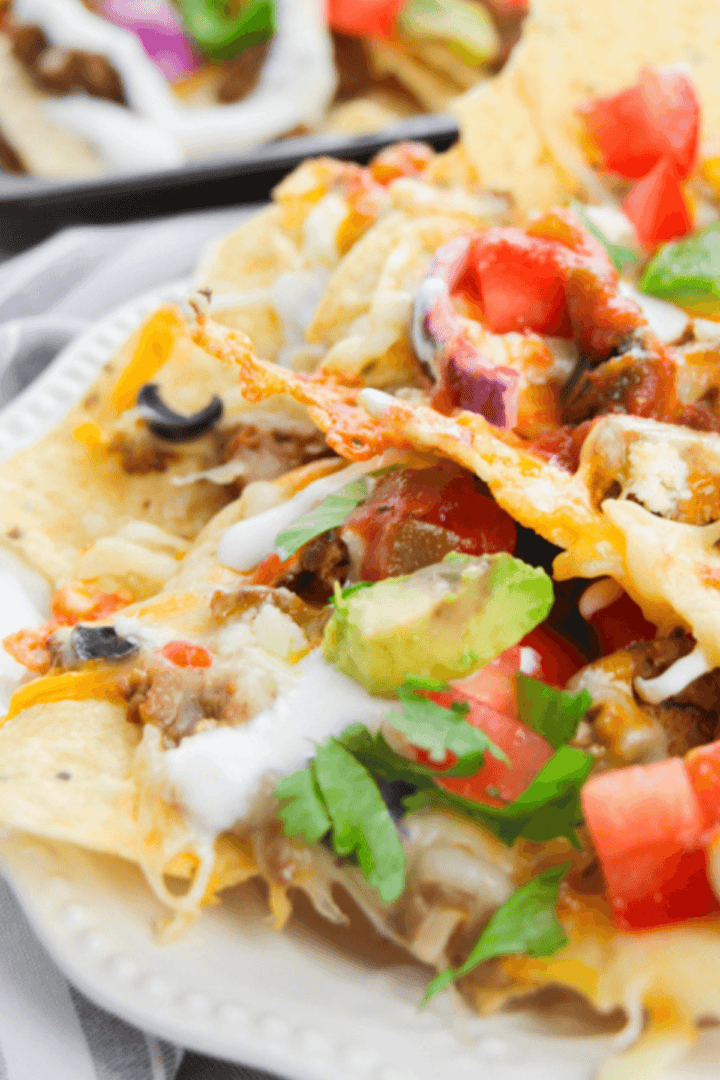 A delicious plate of loaded nachos is generously topped with melted cheese, creamy avocado, juicy tomatoes, spicy jalapeños, zesty onions, and a dollop of tangy sour cream for the ultimate snacking experience.