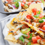A plate of loaded nachos topped with melted cheese, diced tomatoes, sliced jalapeños, and drizzled with sour cream. A similar inviting tray of nachos is visible in the background.