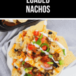 A delicious plate of loaded nachos, brimming with toppings such as tomatoes, onions, olives, lettuce, sour cream, and cheese, rests invitingly on a striped cloth.