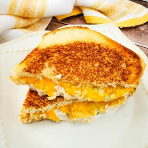 An Air Fryer Tuna Melt grilled cheese sandwich, cut in half, sits on a white plate with a yellow and white striped towel in the background.