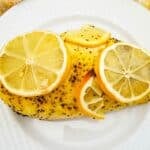 A seasoned Baked Lemon Pepper Chicken breast topped with four lemon slices, served on a white textured plate with a yellow and white towel in the background.