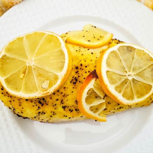 A seasoned Baked Lemon Pepper Chicken breast topped with four lemon slices, served on a white textured plate with a yellow and white towel in the background.