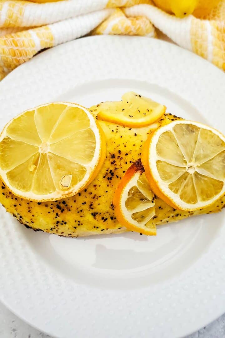 A seasoned Baked Lemon Pepper Chicken breast topped with four lemon slices, served on a white textured plate with a yellow and white towel in the background.