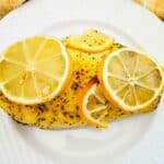 Baked Lemon Pepper Chicken breast seasoned with ground pepper, topped with fresh lemon slices, served on a white plate with a yellow and white towel nearby.