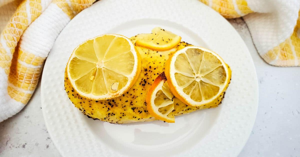 Baked Lemon Pepper Chicken breast seasoned with ground pepper, topped with fresh lemon slices, served on a white plate with a yellow and white towel nearby.