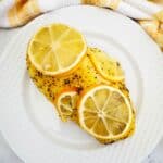 Two breasts, topped with slices , are served on a white plate with a yellow and white towel in the background.