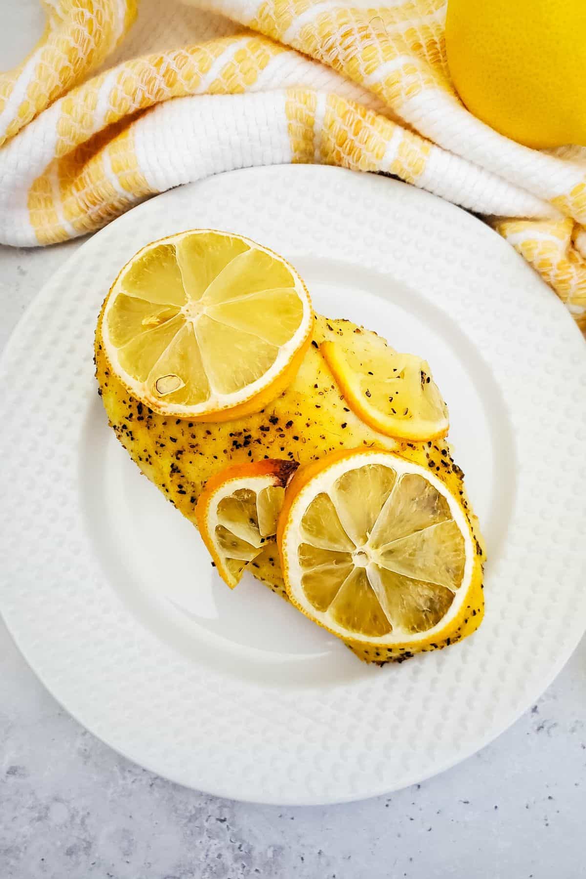 Two breasts, topped with  slices , are served on a white plate with a yellow and white towel in the background.