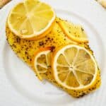 Baked Lemon Pepper Chicken breast topped with fresh lemon slices, served on a white plate with a yellow and white towel in the background.