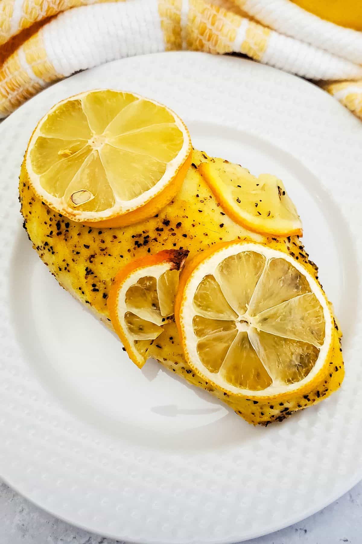 Baked Lemon Pepper Chicken breast topped with fresh lemon slices, served on a white plate with a yellow and white towel in the background.