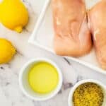 Two raw breasts on a white plate, two lemons, a small bowl of olive oil, and a bowl of seasoning sit on a marble countertop.