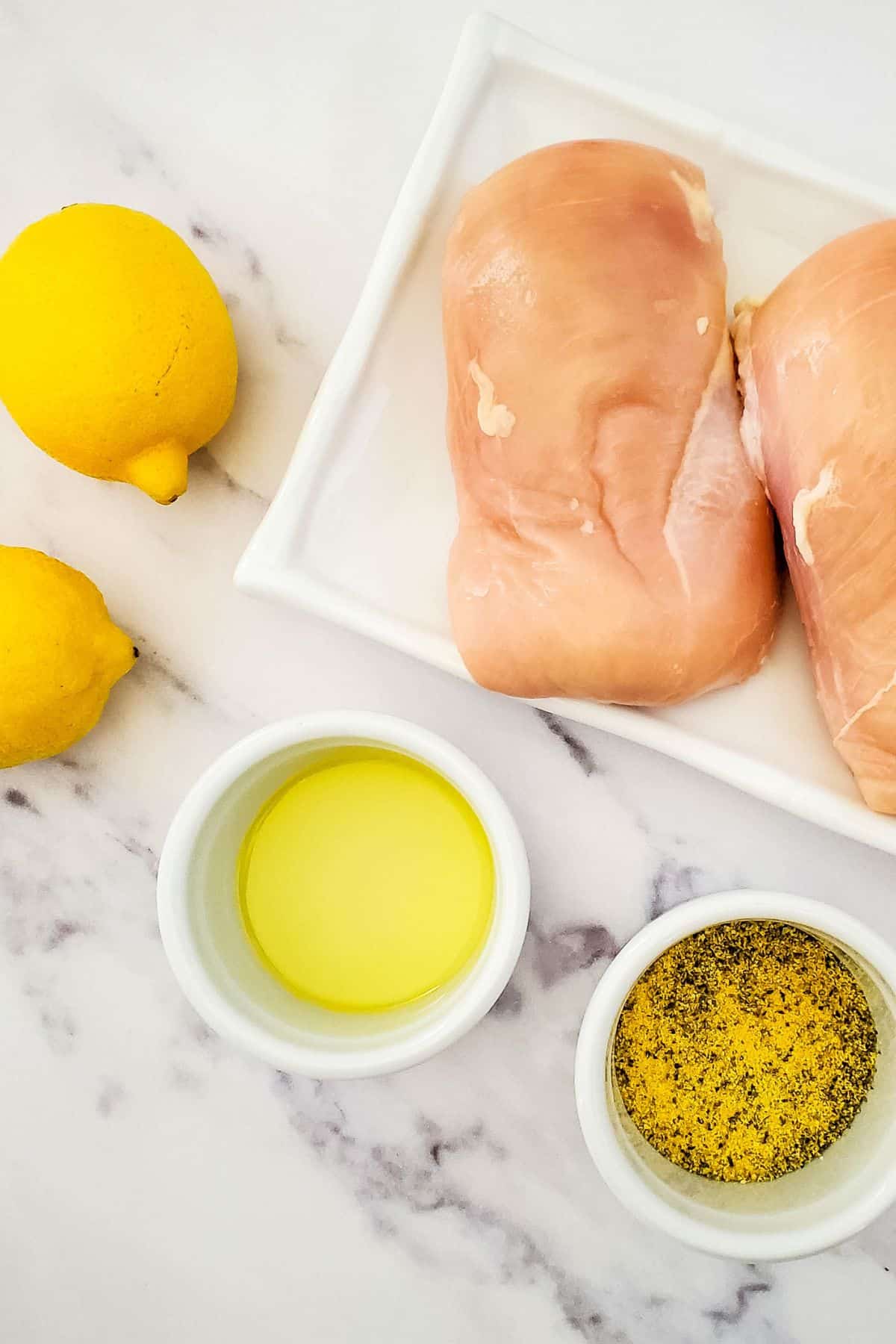 Two raw breasts on a white plate, two lemons, a small bowl of olive oil, and a bowl of seasoning sit on a marble countertop.