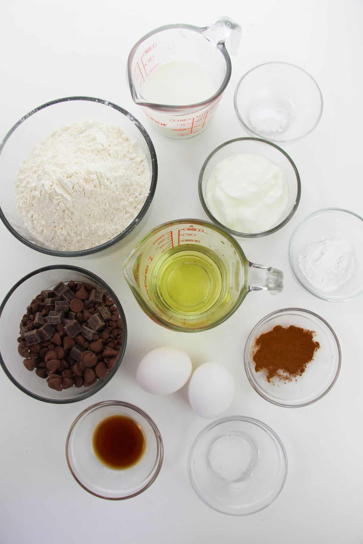 Various baking ingredients are arranged in bowls and measuring cups, ready to create delicious treat featuring flour, sugar, yogurt, eggs, chocolate chips, vanilla, oil, cinnamon, and baking powder.