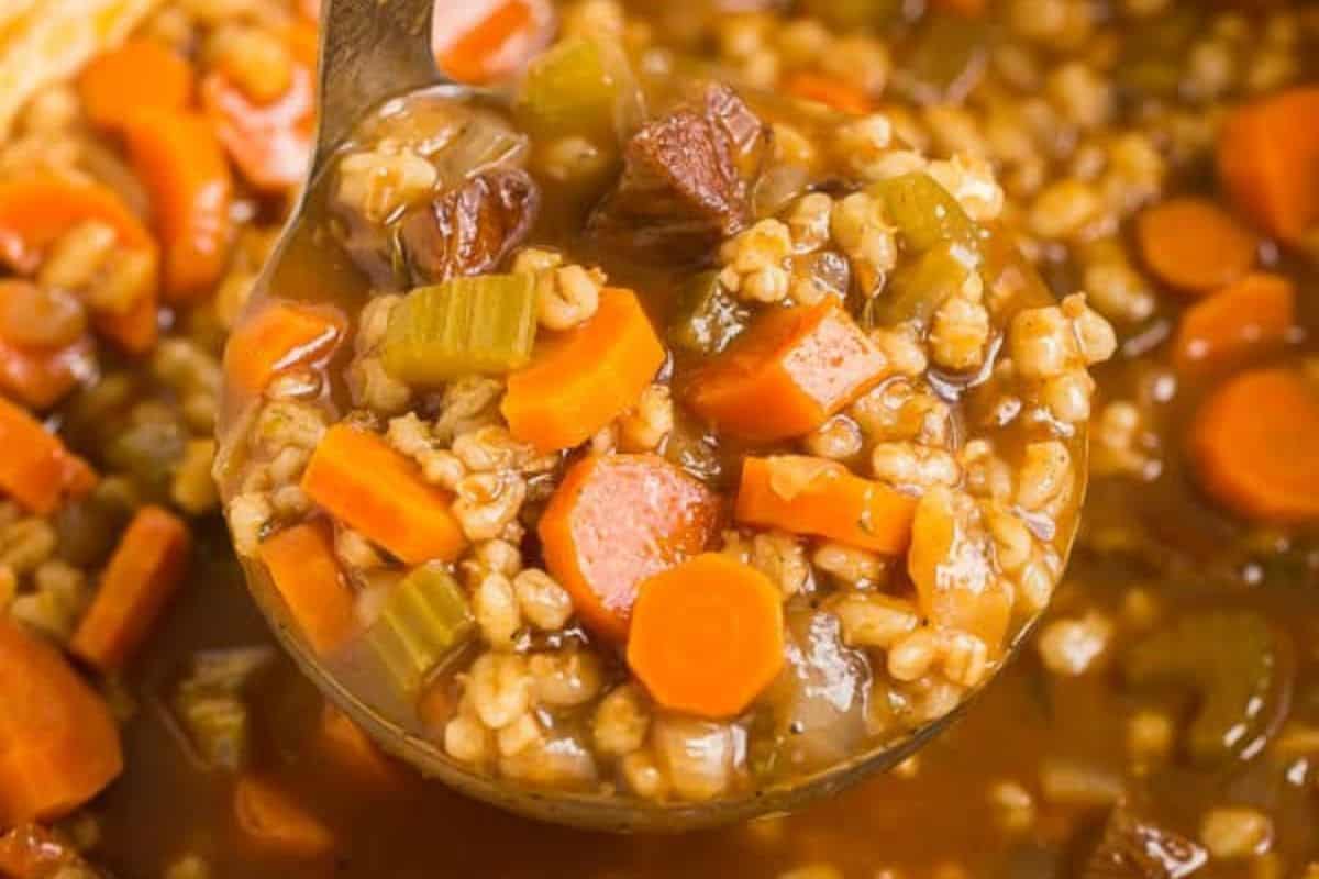 A ladle holds vegetable and barley soup with visible carrots, celery, barley, and chunks of meat in a brown broth—perfect for new mom freezer meals.