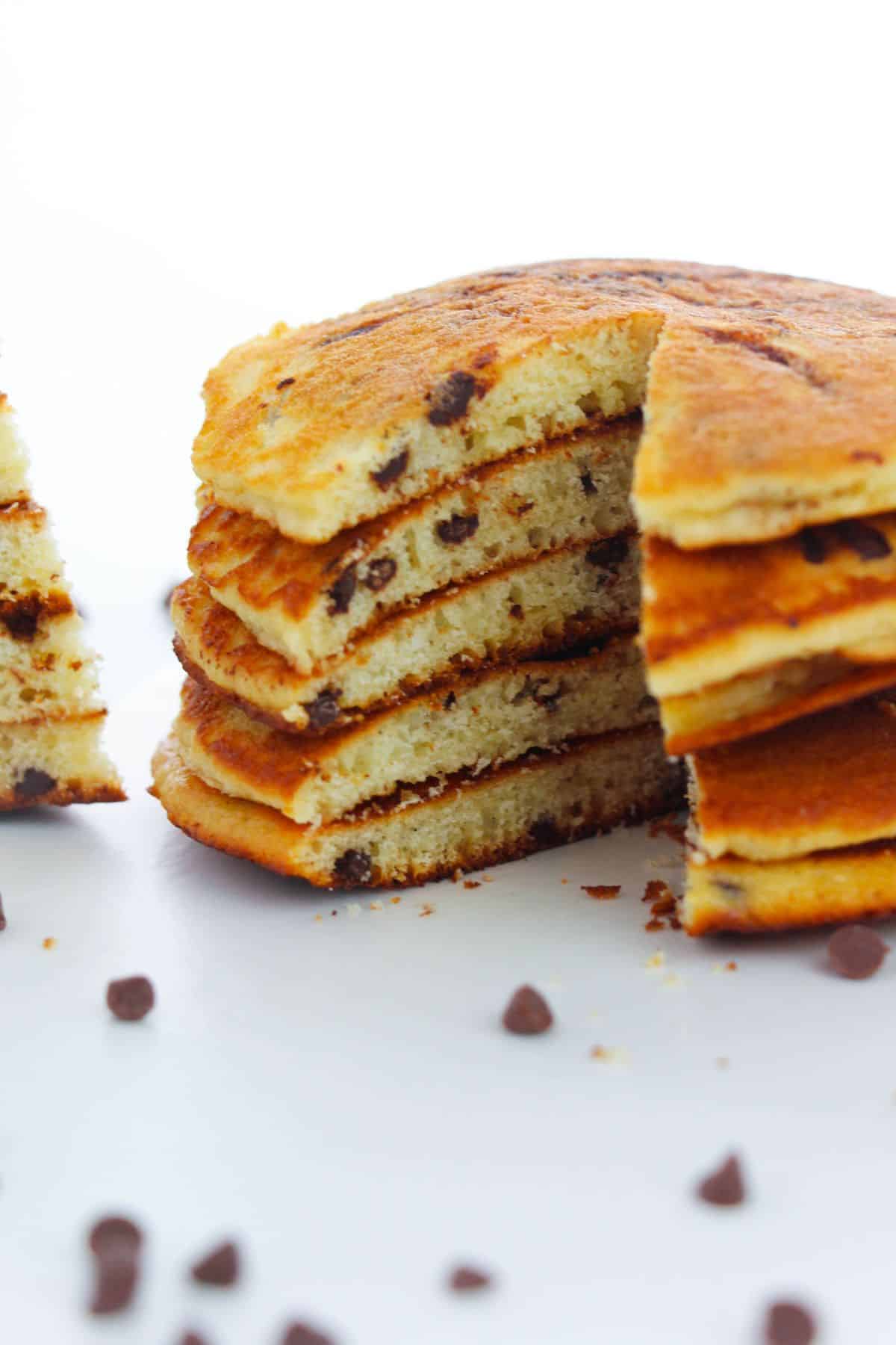 A stack of chocolate chip Cake Mix Pancakes with a wedge cut out, sitting on a white surface with scattered chocolate chips nearby.