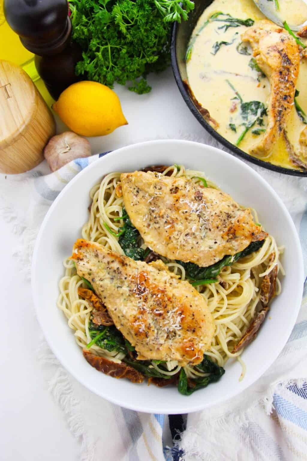 A bowl of Creamy Garlic and Chicken Pasta topped with seasoned chicken breast, sautéed spinach, and grated cheese, served with a creamy sauce, lemon, and herbs in the background.