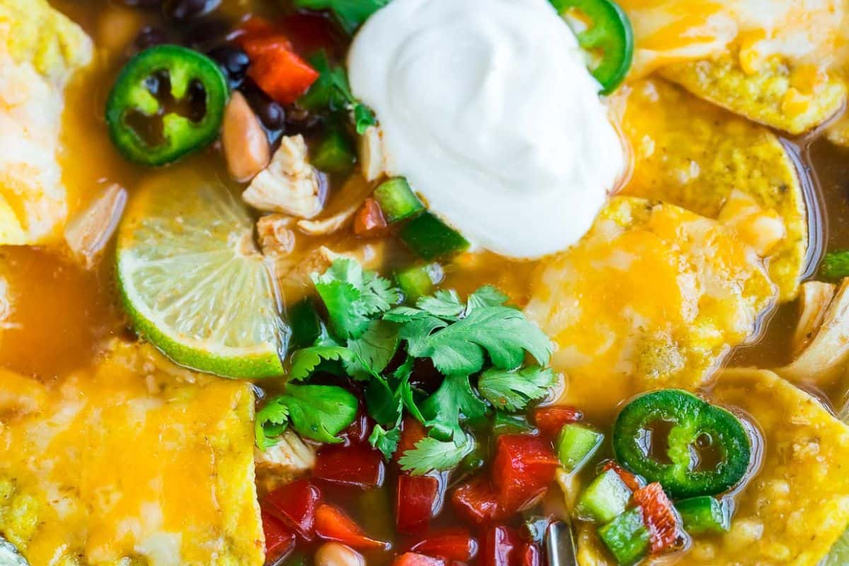A close-up of a bowl of tortilla soup topped with melted cheese, jalapeño slices, black beans, red bell peppers, cilantro, lime, and a dollop of sour cream—perfect for new mom freezer meals.