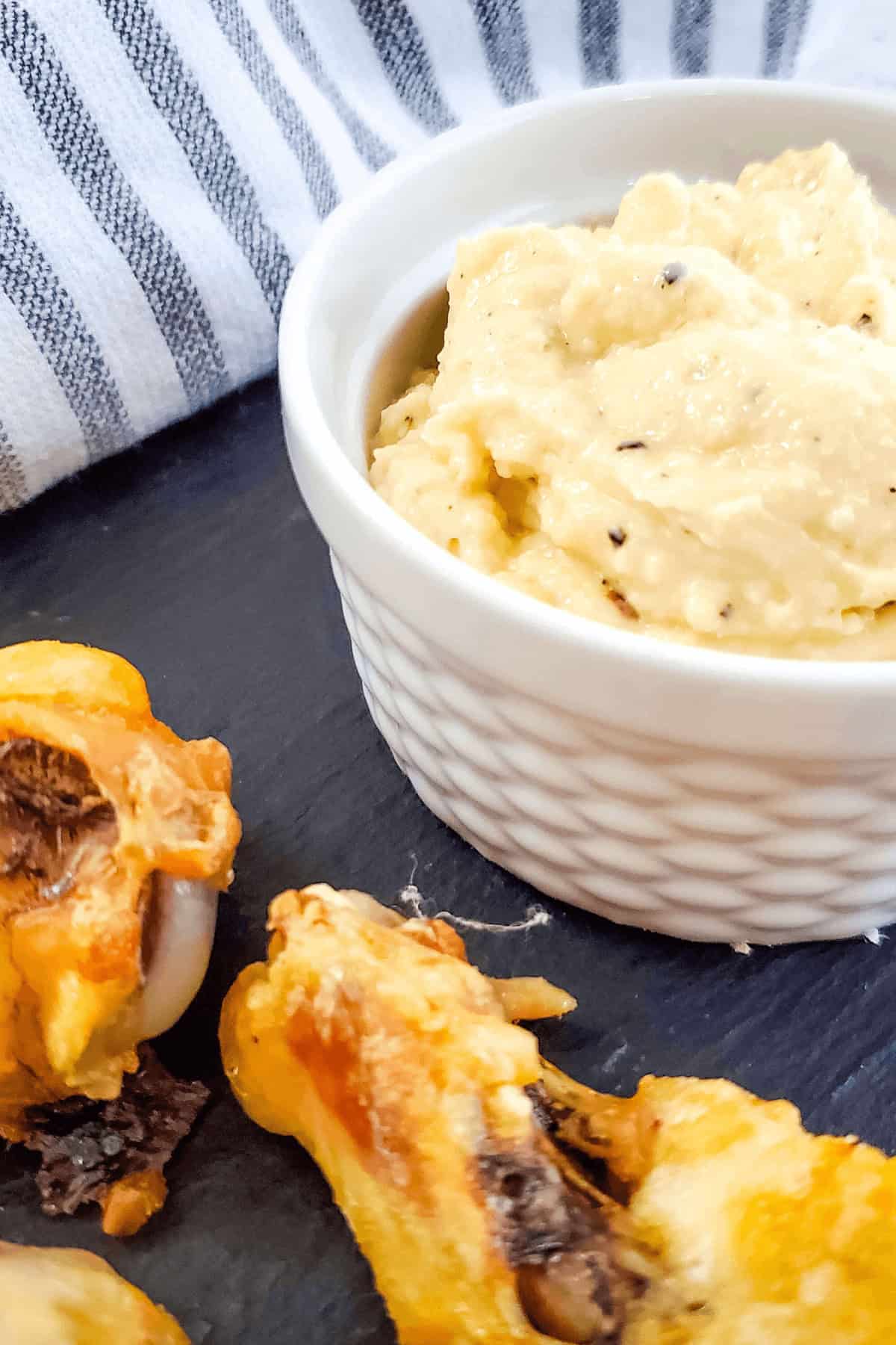 A small bowl of creamy garlic dip on a slate surface, perfectly paired with crunchy orange and brown snacks. The scene hints at a feast of Garlic Parmesan Chicken Wings, while a striped cloth adds a touch of warmth in the background.