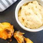 A bowl of creamy dip sits next to fried Garlic Parmesan Chicken Wings on a slate serving board, with a striped cloth nearby.