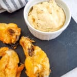 Chicken drumsticks and a bowl of creamy garlic Parmesan dip grace a slate serving board, with a striped cloth in the background.
