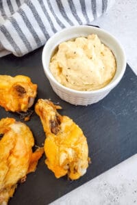 Chicken drumsticks and a bowl of creamy garlic Parmesan dip grace a slate serving board, with a striped cloth in the background.