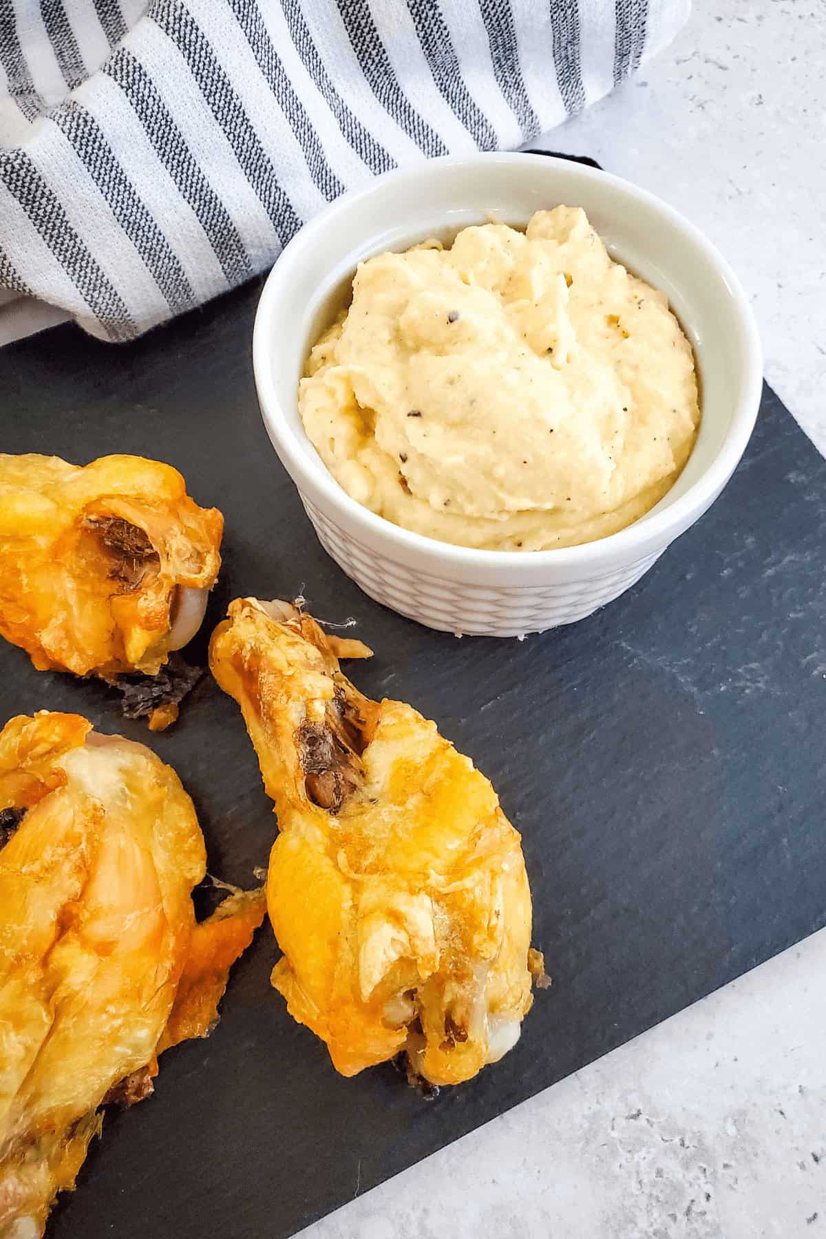 Chicken drumsticks and a bowl of creamy garlic Parmesan dip grace a slate serving board, with a striped cloth in the background.