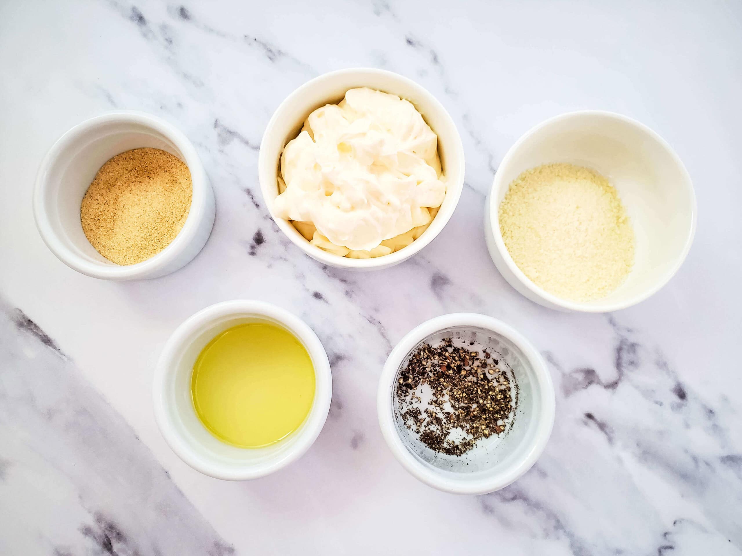 Five white bowls on a marble surface hold ingredients essential for Garlic Parmesan Chicken Wings: garlic powder, mayonnaise, grated parmesan, olive oil, and a salt-pepper mix.