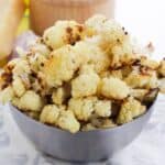A metal bowl filled with garlic roasted cauliflower florets sits on a patterned cloth, with a wedge of cheese and other ingredients blurred in the background.