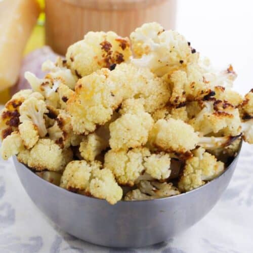 A metal bowl filled with garlic roasted cauliflower florets sits on a patterned cloth, with a wedge of cheese and other ingredients blurred in the background.
