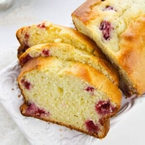 A loaf of Lemon Berry Loaf, partially sliced to reveal fresh raspberries inside, is displayed on a white plate.