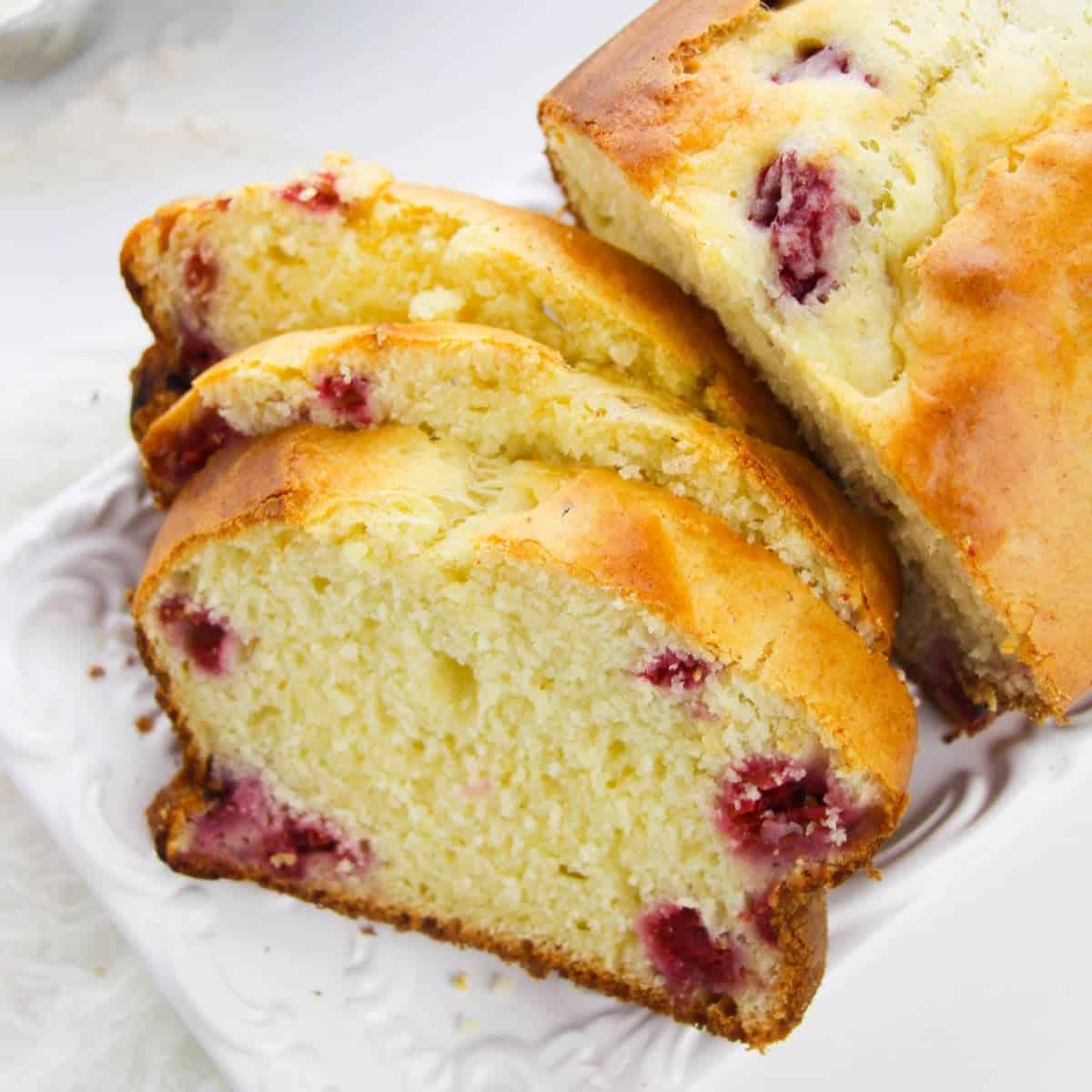 A loaf of Lemon Berry Loaf, partially sliced to reveal fresh raspberries inside, is displayed on a white plate.