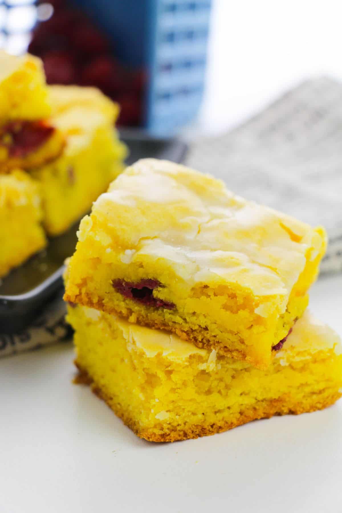 Two pieces of Lemon Blondies with a light glaze and a layer of fruit filling are stacked on a white surface, with more cake pieces on a plate in the background.