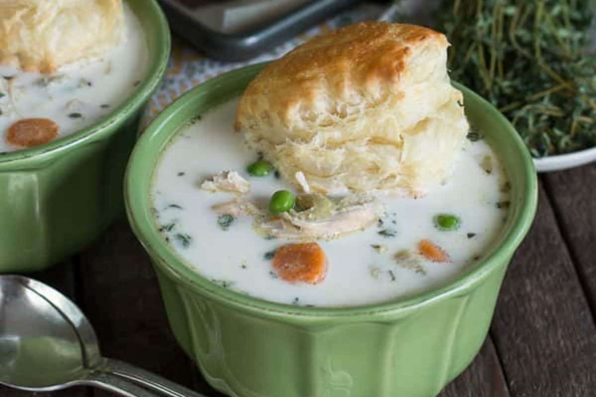 A green bowl filled with creamy soup containing vegetables and chicken, topped with a golden biscuit—perfect as one of those comforting new mom freezer meals; a spoon and some herbs are nearby.