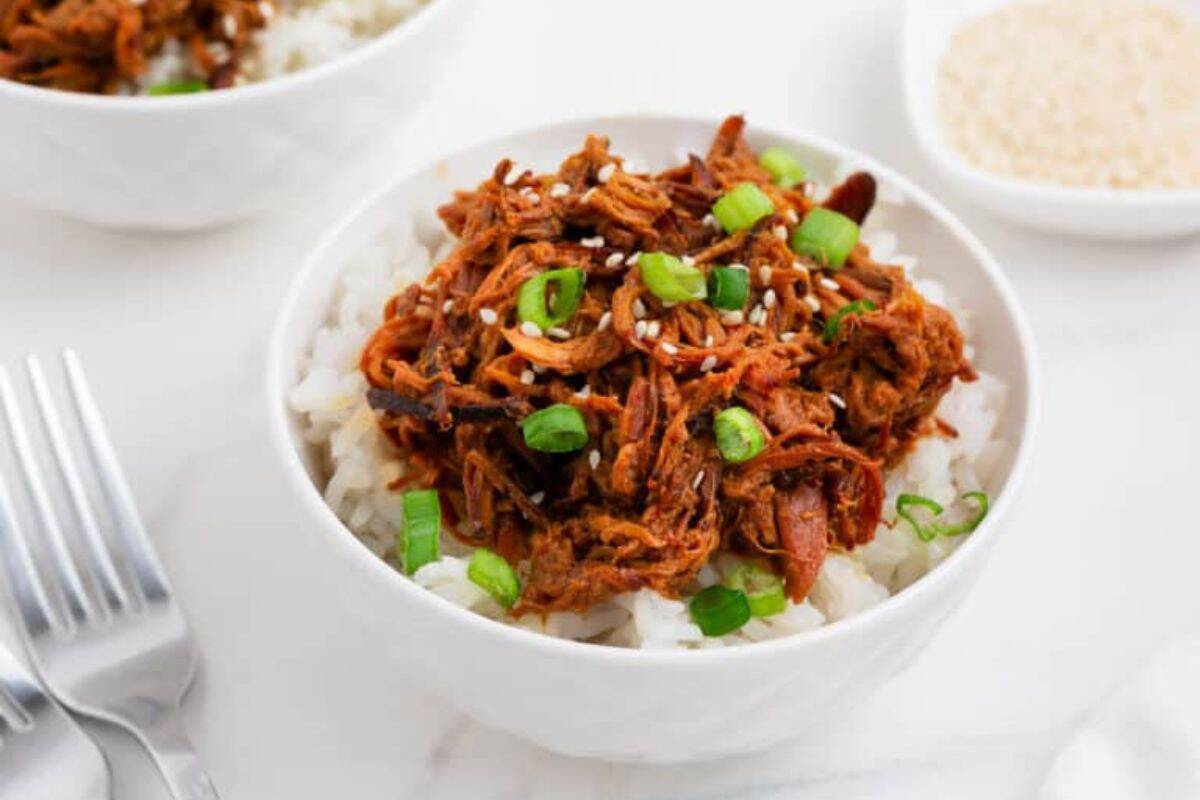 A white bowl filled with white rice topped with shredded chicken, sliced green onions, and sesame seeds—an easy and nourishing option for new mom freezer meals—served alongside a bowl of sesame seeds and forks.